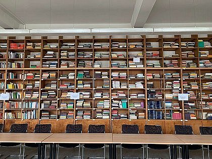 Picture of the gender studies library. It shows a full bookshelf reaching up to the ceiling, with tables and chairs in front of it that can be used as workstations.